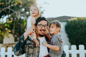 5 Ways to Involve Kids in the Family Budget. A father is smiling and holding his small son with his left hand and his smaller daughter with his right hand as she is sitting behind on his neck and behind his back