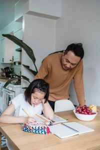 How to make it a budget-friendly March Break. A father is standing next to his daughter while she is writing in a book on the kitchen table